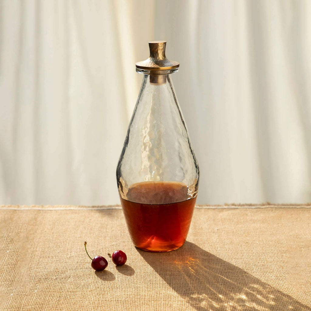Handcrafted pebbled glass decanter with hammered aluminum stopper, filled with rich liquid, on a textured surface.